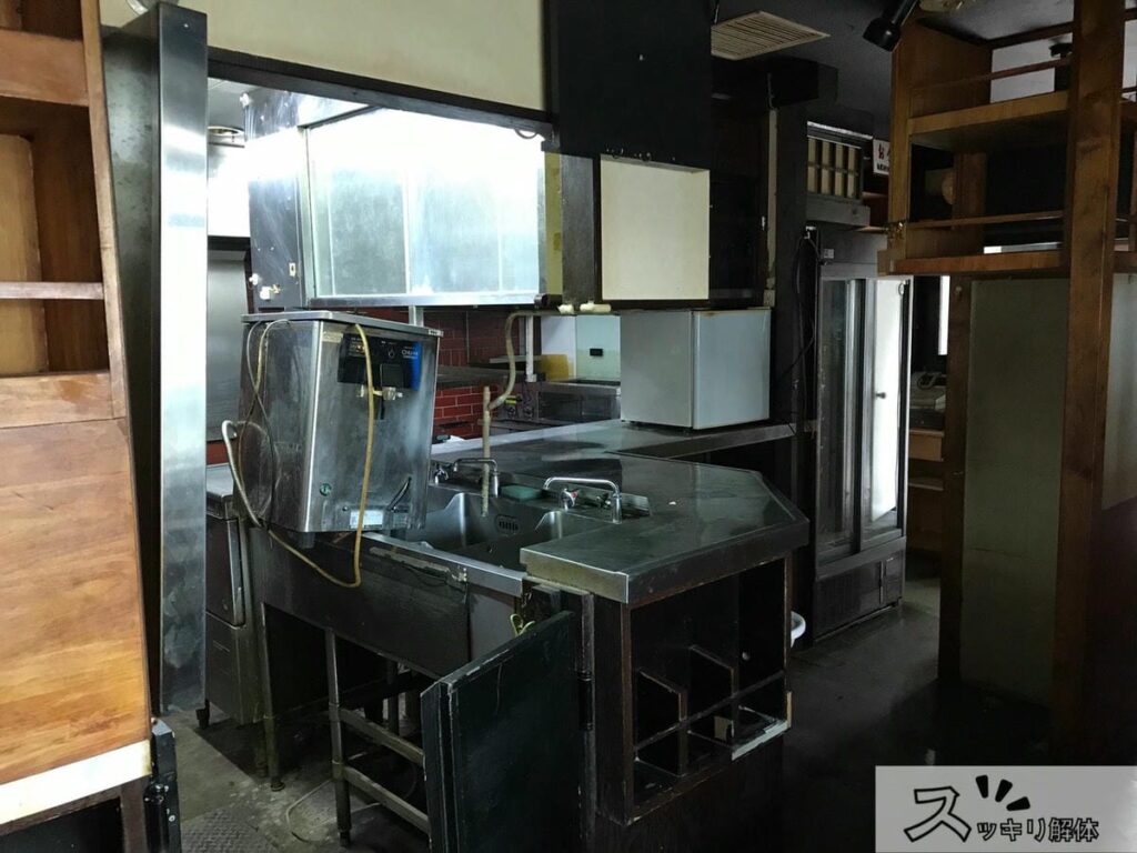 ステンレス製のシンクやカウンター、棚などの内装設備がそのまま残っている、解体工事前の飲食店の厨房