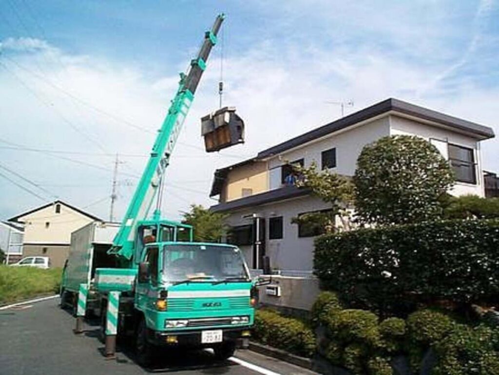 住宅街でクレーン車を使い、戸建て住宅の2階窓からピアノを吊り下げて搬出している特殊作業の様子