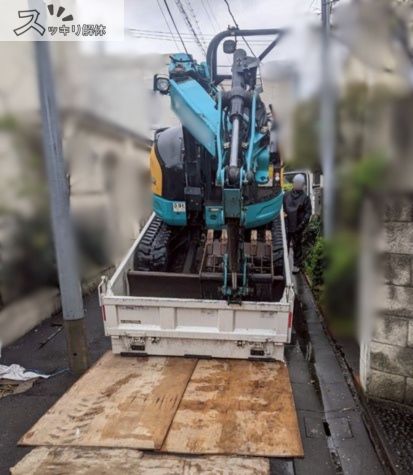 住宅に囲まれた狭い道路で小型のショベルカーをトラックの荷台へ積み込む様子。道路を保護するため、トラックの後方道路に木の板を敷いている。