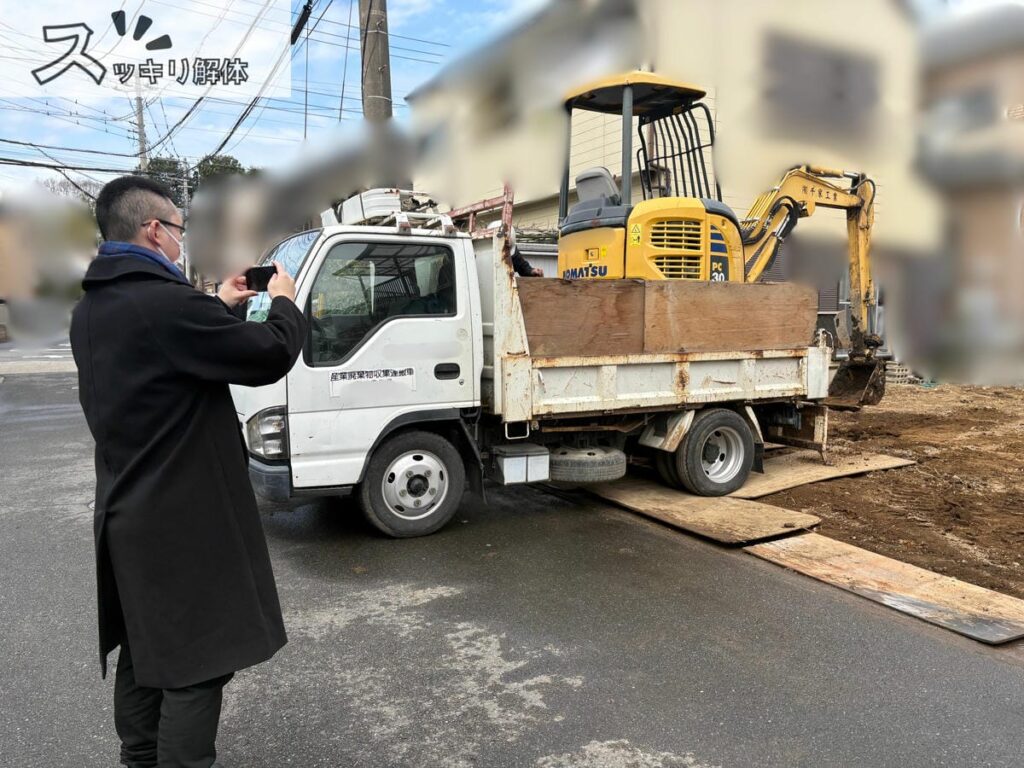 解体工事が終わり更地になった現場の前で、トラックの荷台に積み込まれた黄色いミニショベルをスマホで撮影している様子。