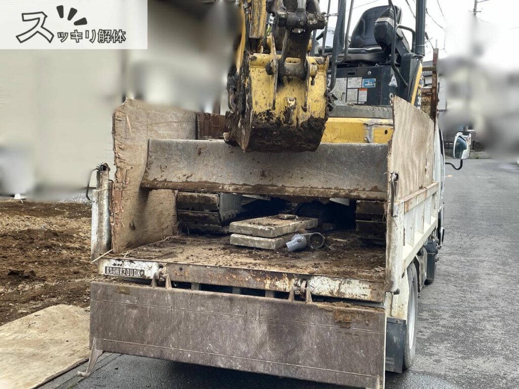 ミニショベルが積み込まれたトラックの荷台を後方から見た様子。荷台には木の板で囲いがされており、トラック後方のあおりは開いている。