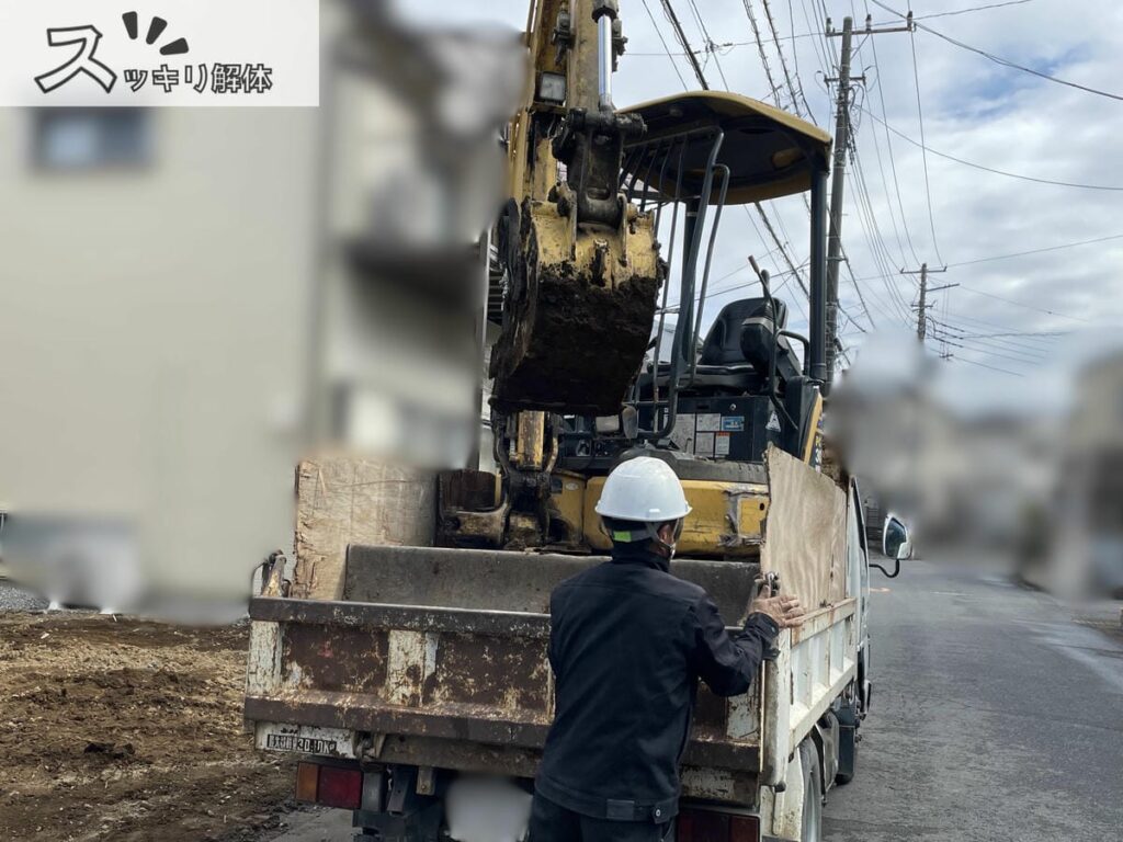 更地になった解体現場の前で、ミニショベルを積んだトラックの後部に立ち、ヘルメット姿の作業員が荷台後方のあおりを手でしっかりと固定している様子。