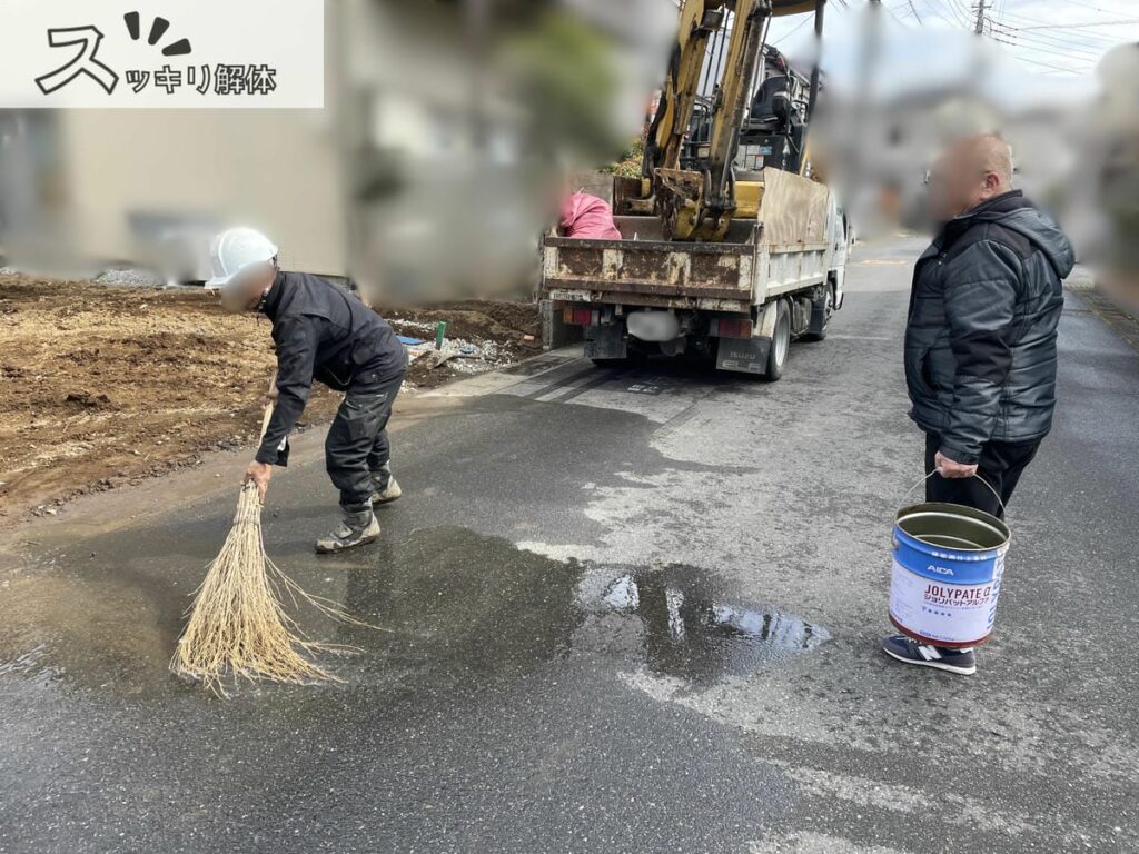 ミニショベルが積まれたダンプトラックの後方で、土で汚れた道路を作業員2人が掃除している様子。右側の1人は水撒きに使ったバケツを持ち、左の1人は竹ぼうきで土汚れを掃いている。